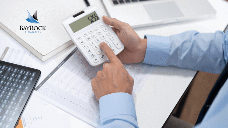 A person in a blue shirt uses a calculator at a desk with financial documents, a laptop, and a tablet. The BayRock Financial logo is visible in the top left corner.