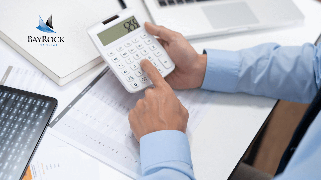 A person in a blue shirt uses a calculator at a desk with financial documents, a laptop, and a tablet. The BayRock Financial logo is visible in the top left corner.