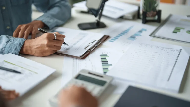 A person writes on a clipboard surrounded by financial documents, charts, and a calculator on a desk, suggesting analysis or business planning.