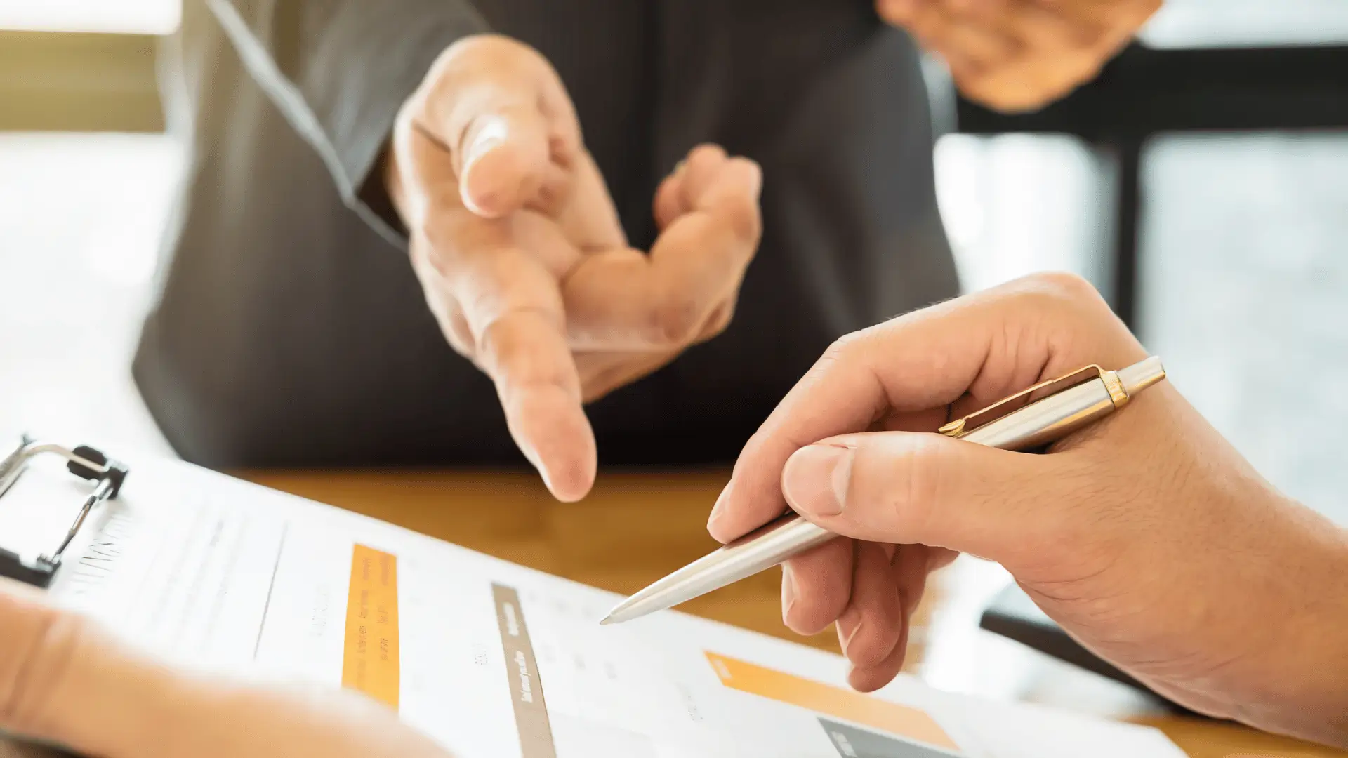 Close-up of two people in business attire discussing a document on a clipboard. One person holds a pen over the paper, while the other gestures with open hands, suggesting a discussion or negotiation.